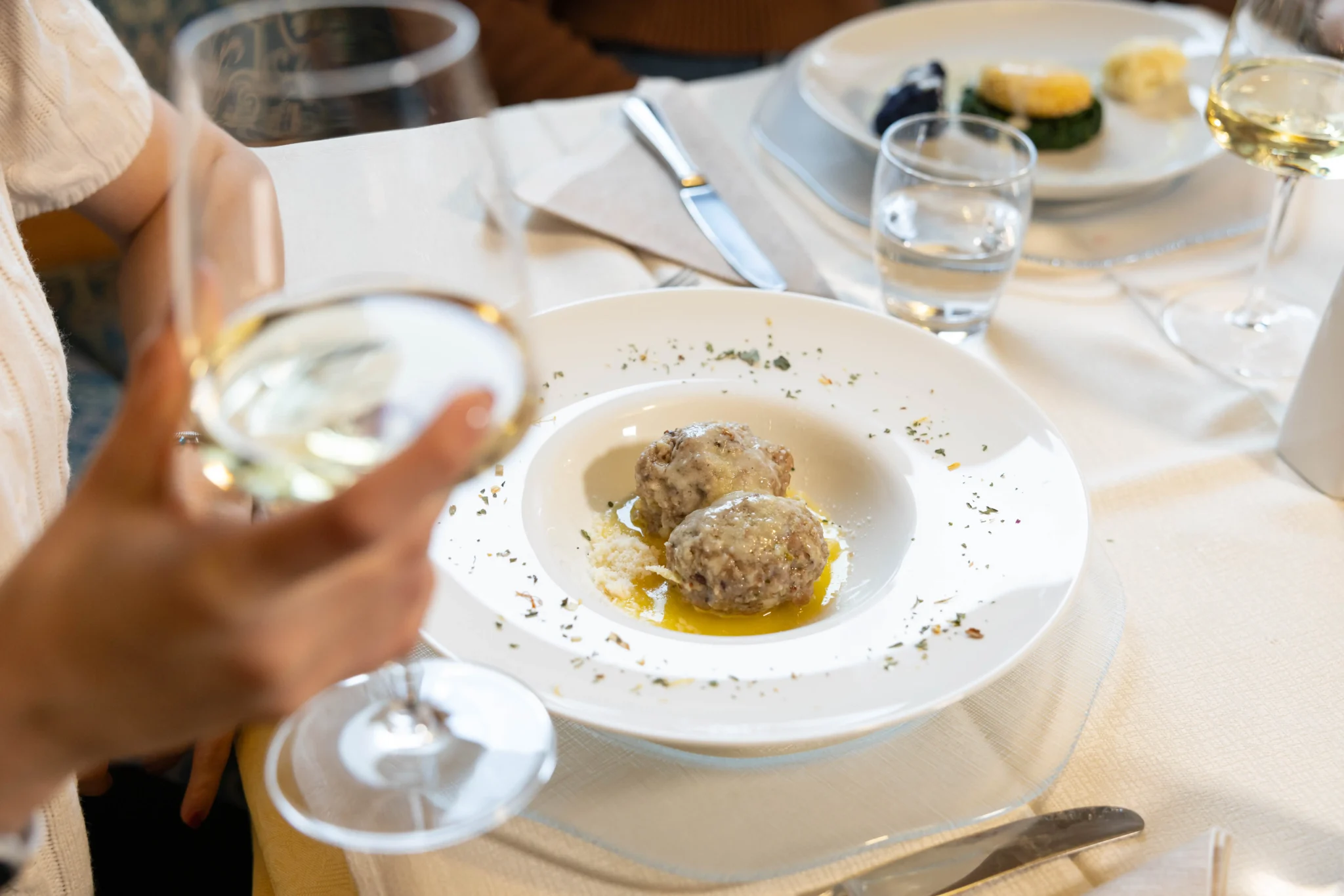 Piatto di canederli con salsa e calice di vino bianco su tavola imbandita.