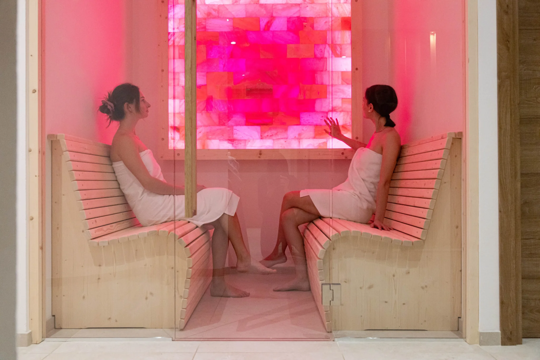 Due donne in accappatoio siedono in una sauna con luci rosa.