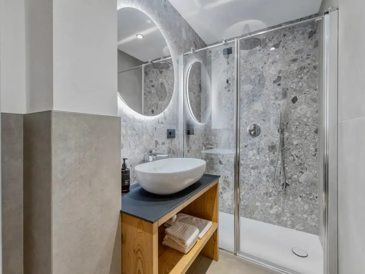 Moderno bagno con doccia in vetro, lavabo su mobile in legno e specchio rotondo retroilluminato.