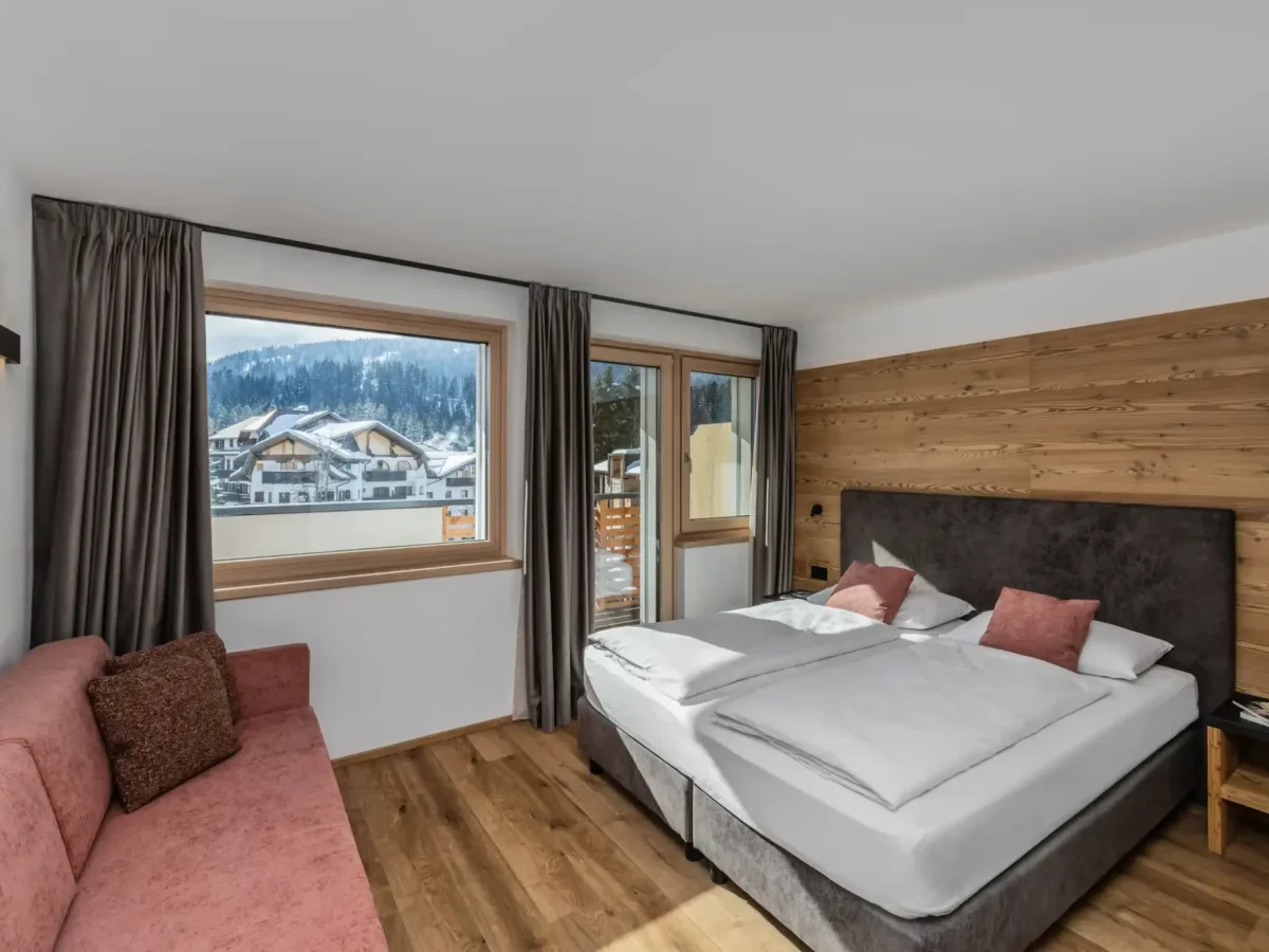 Camera d'albergo moderna con letto matrimoniale, divano e vista panoramica sulle montagne innevate.
