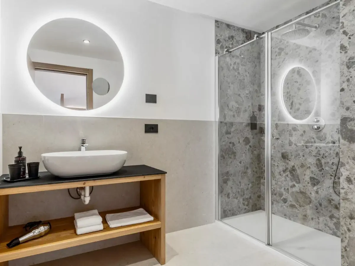 Bagno moderno con lavandino, specchio rotondo illuminato e doccia con parete in pietra.