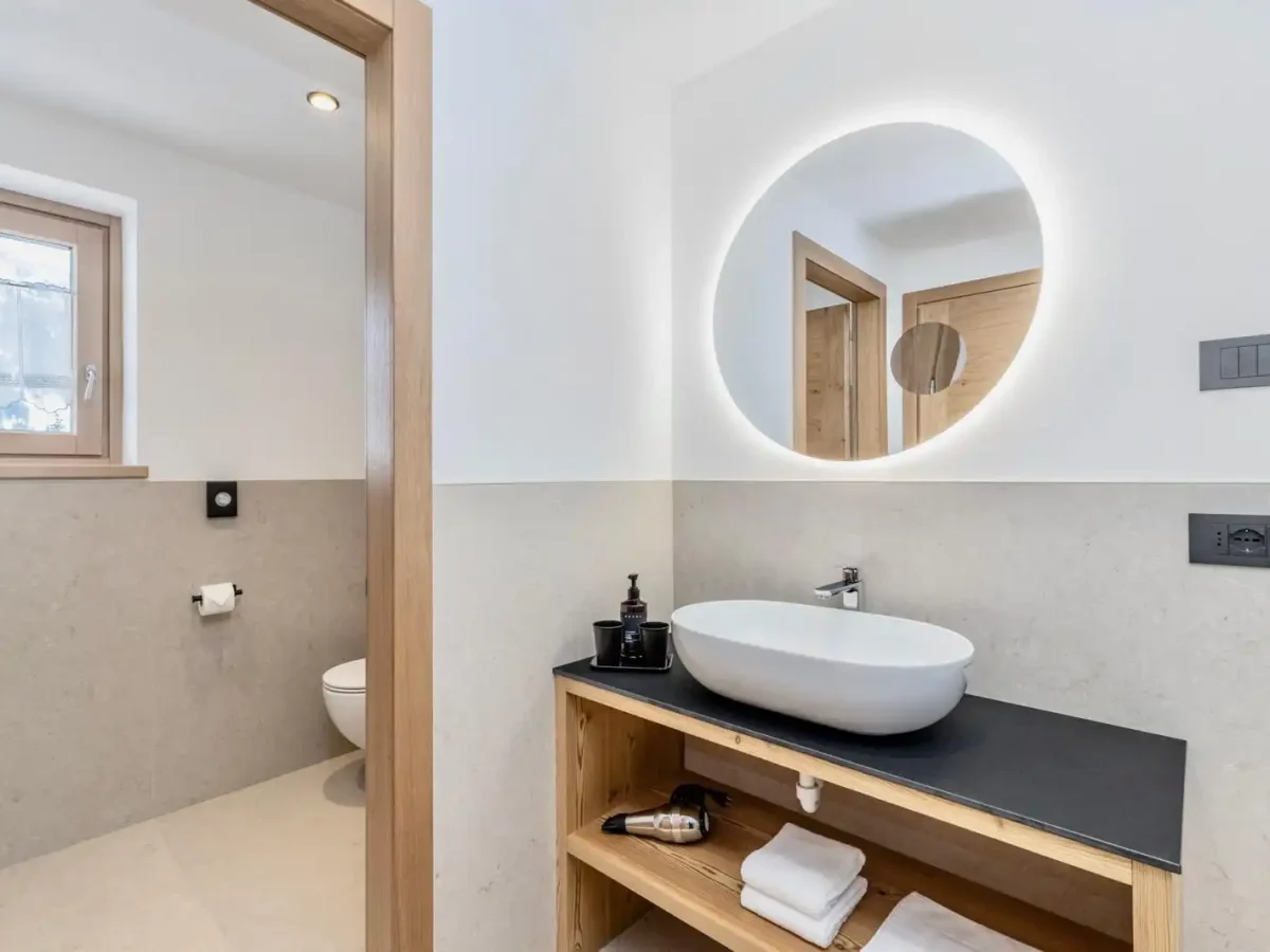 Bagno moderno con lavabo elegante, specchio rotondo illuminato e dettagli in legno.
