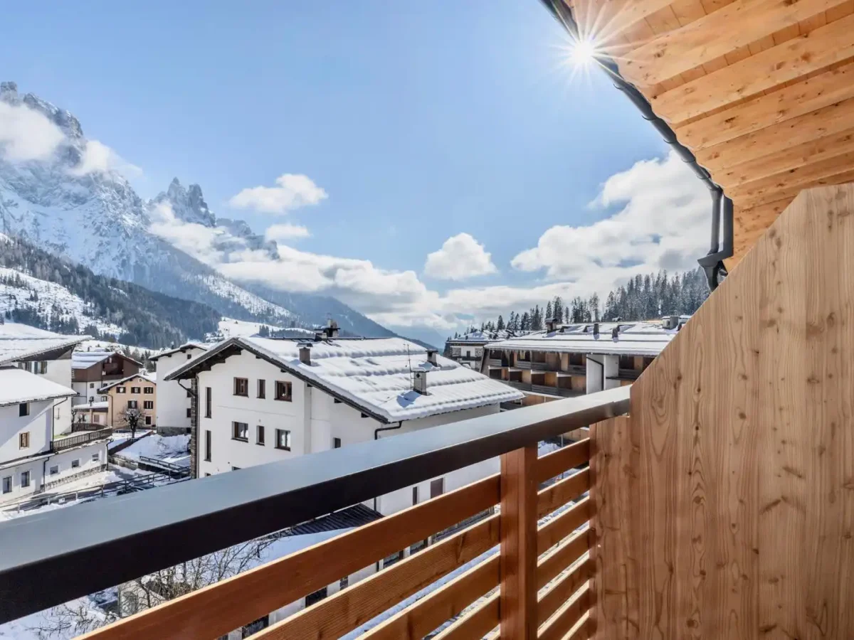Vista panoramica di un villaggio di montagna innevato con montagne sullo sfondo e un balcone in legno in primo piano.