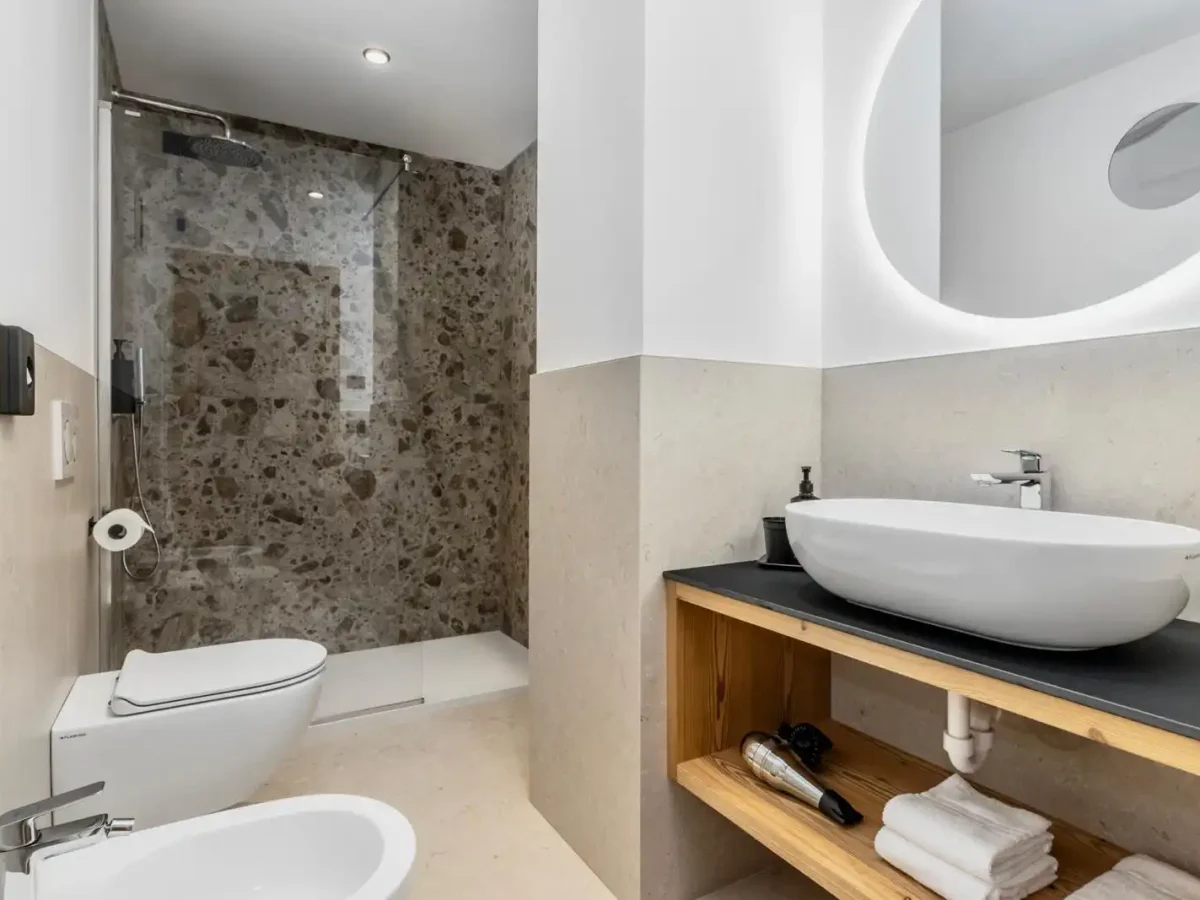 Bagno moderno con doccia in pietra, lavabo ovale, e specchio illuminato.