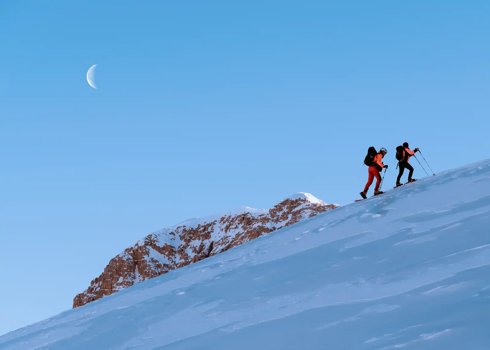 Due persone fanno scialpinismo su un pendio innevato sotto una luna crescente.